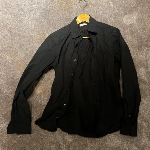 Svart skjorta från Burberry - Säljer en stilren svart skjorta från Burberry i mycket bra skick. Skjortan har långa ärmar och knappar framtill. Perfekt för både vardag och festliga tillfällen. Passar bra till både jeans och kostymbyxor. Ett måste i garderoben för den som gillar klassisk stil! 🖤