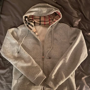 Grå burberry hoodie - Säljer en snygg grå hoodie från Burberry med klassiskt rutigt mönster i luvan. Den har dragkedja framtill och är i mycket bra skick. Perfekt för höst och vår! Skriv för frågor! 