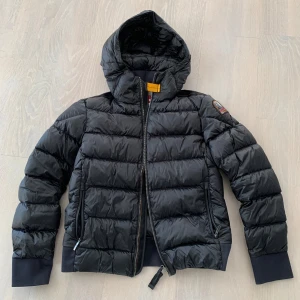 Svart dunjacka från Parajumpers - Säljer en snygg svart dunjacka från Parajumpers, modell Sheen. Jackan har en cool patch på ärmen och en praktisk huva. Perfekt för kyliga dagar med sin quiltade design och dragkedja framtill. Passar både till vardags och lite finare tillfällen. 🖤