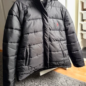 Svart dunjacka från Ralph Lauren - Säljer en snygg svart dunjacka från Ralph Lauren. Den har en liten röd logga på bröstet. Den har dragkedja framtill och två praktiska fickor. Den är inte äkta, som man ser på loggan, det står att storleken är s, men den är s/m. Den är helt oanvänd.