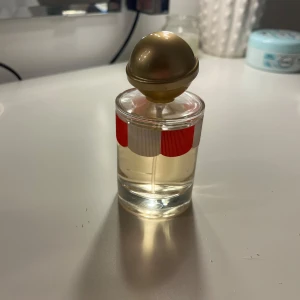 Parfym - Säljer en elegant parfymflaska med en unik röd och vit randig design. Flaskan har en rund, gyllene kork som ger en lyxig känsla. Flaskan är genomskinlig så att du kan se den ljusa vätskan inuti.