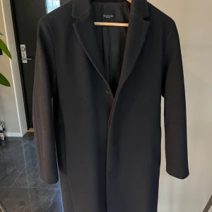 Svart kappa från Selected - Säljer en elegant marinblå rock från Selected i mycket bra skick. Den har en klassisk design med knappar framtill och är perfekt för höst och vinter. Kappan är långärmad och har en stilren passform som passar både till vardags och fest. Fodret är mjukt och bekvämt.