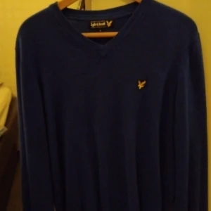 Blå tröja från Lyle & Scott - Säljer en snygg blå tröja från Lyle & Scott med deras ikoniska logga på bröstet. Tröjan har en v-ringad design och långa ärmar, perfekt för både vardag och lite finare tillfällen. Den är i bra skick och passar perfekt till höst- och vårgarderoben.