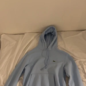 Ljusblå Lacoste Hodie. - Säljer en Lacoste Hoodie i storlek XS. Ljusblå färg med Lacoste loggan på bröstet. Använd en gång. Bättre pris vid snabb affär. Funkar för alla årstider! Nypris 1750 kr.