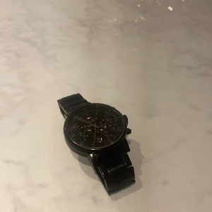 Svart klocka från Tommy Hilfiger - Säljer en stilren svart klocka från Tommy Hilfiger. Den har en svart urtavla med tydliga siffror och tre mindre urtavlor för extra funktioner. Armbandet är också svart och ger en elegant look. Perfekt för både vardag och fest!