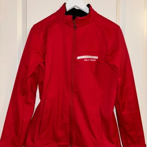 Röd jacka från Helly Hansen - Säljer en snygg röd jacka från Helly Hansen. Den har en dragkedja framtill och en praktisk bröstficka med dragkedja. Perfekt för höst och vår, och den är i mycket bra skick. Passar både till vardags och lite sportigare aktiviteter. Storlek M