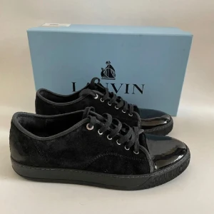 Svarta sneakers från Lanvin - Snygga svarta sneakers från Lanvin i både mocka och lack. De har en stilren design med snörning och en glansig tå. Perfekta för både vardag och fest! Skorna kommer i originalkartong.