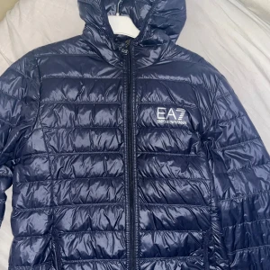 Mörkblå pufferjacka från Emporio Armani - Säljer en snygg mörkblå pufferjacka från Emporio Armani. Jackan har en glansig finish och är perfekt för kyliga dagar. Den har en dragkedja framtill och en huva för extra skydd. EA7-loggan syns tydligt på bröstet. Perfekt för både vardag och lite finare tillfällen!