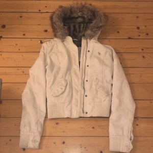 Vit jacka med pälsfodrad huva från Abercrombie & Fitch - Säljer en snygg vit jacka från Abercrombie & Fitch i storlek S/M. Den har en mysig pälsfodrad huva och ribbade muddar. Perfekt för kyliga dagar! Jackan har en dragkedja och knappar framtill för extra värme.