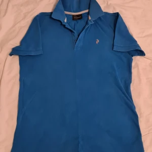 Sommar25. Blå pikétröja från Peak Performance M - Säljer en snygg blå pikétröja från Peak Performance. Perfekt för både vardag och sportiga aktiviteter. Tröjan har en klassisk krage och korta ärmar, vilket gör den idealisk för sommardagar. Den är i bra skick och redo för nya äventyr!