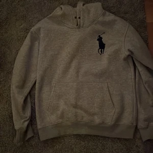 Grå hoodie från Ralph Lauren - Säljer en snygg grå hoodie från Ralph Lauren med den klassiska polospelaren broderad på bröstet. Tröjan har en stor ficka fram och en cool nummerdetalj på ärmen. Perfekt för en avslappnad stil eller en kylig dag. Passar både till vardags och chillkvällar. Storlek S