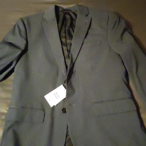 Blå Grå kavaj från H&M - Säljer en snygg blå grå kavaj från H&M i slim fit med stretch. Den är i nytt skick och gjord av återvunnen polyester, vilket är toppen för miljön! Perfekt för både fest och mer formella tillfällen. Storleken är herr M. Passar bra till både höst och vår.