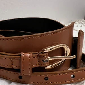Brunt skärp med guldfärgat spänne - Säljer ett snyggt brunt skärp i skinn med ett elegant guldfärgat spänne. Perfekt för att ge din outfit en stilren touch. Skärpet har flera hål för justerbar passform och är i nytt skick. Passar perfekt till både vardag och fest!