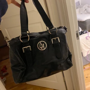 Svart handväska från Armani Jeans - Säljer en snygg svart handväska från Armani Jeans i mycket bra skick. Väskan har ett stilrent märke framtill och är utrustad med dragkedja och justerbara remmar med spännen. Perfekt för både vardag och fest!