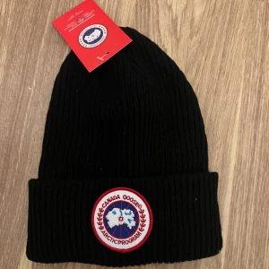 Svart ribbad mössa från Canada Goose - Säljer en snygg svart ribbad mössa från Canada Goose. Den har en broderad logga framtill och är perfekt för kalla dagar. Mössan är i nytt skick och kommer med originaltaggen kvar. Perfekt för vintern! 🖤