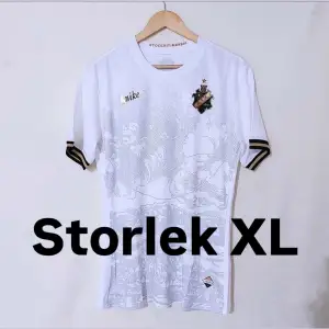 Säljer en snygg vit fotbollströja från Nike i storlek XL. Tröjan har korta ärmar med svarta och guldiga detaljer på ärmsluten. Den har ett subtilt mönster på framsidan och en klubbemblem på bröstet. Perfekt för fotbollsfans eller som en stilren vardagströja!