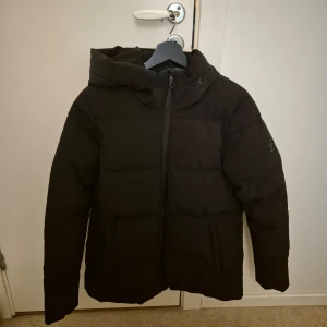 Svart vinterjacka från Cross Sportswear - Säljer en svart dunjacka från Cross Sportswear i storlek M. Jackan är perfekt för kalla dagar med sin pufferstil och långa ärmar. Den har en dragkedja framtill och en logga på ärmen.