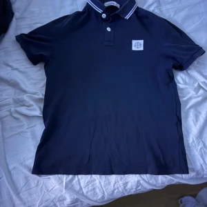 Mörkblå pikétröja med logga - Säljer en snygg mörkblå pikétröja från Stone Island. Den har en klassisk krage med vita ränder och knappar framtill. Tröjan är kortärmad och har en broderad logga på bröstet. Perfekt för både vardag och fest! Bra skick nypris 2299 mitt pris 529