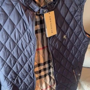 Mörkblå quiltad väst från Burberry - Säljer en snygg mörkblå quiltad väst från Burberry i nytt skick. Västen har ett klassiskt rutigt mönster på insidan och en dragkedja framtill. Perfekt för höst och vår, och den har en stilren look med en liten broderad logga på bröstet. Passar både till vardags och fest!