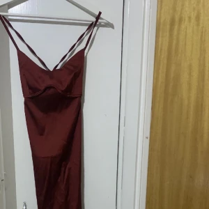 Vinröd långklänning i satin - Säljer en elegant vinröd långklänning i satin med smala axelband och öppen rygg. Klänningen har en tight passform och är perfekt för festliga tillfällen. Den har en vacker glans och knytning i ryggen för en extra detalj. Passar perfekt till speciella tillfällen.
