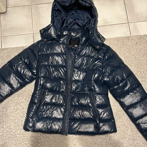 Mörkblå glansig pufferjacka med huva - Säljer en snygg mörkblå  pufferjacka i glansigt material. Jackan har en praktisk huva och dragkedja framtill. Perfekt för kyliga dagar och håller dig varm och stilren. Passar både till vardags och lite finare tillfällen. 💙helt oanvänd💞