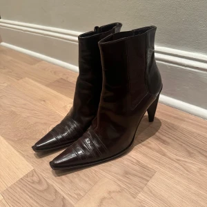 Mörkbruna ankelstövlar i lackläder - Väldigt snygga ankelstövlar med klack + spetsig tå från Italien!  Storlek: Ca 38-39 (39 framgår på baksidan, men jag är 38-39) Klackar: Ca 10 cm Färg: Mörkbrun Material: Lackläder/blankt skinn Skick: Mycket bra