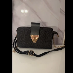 Svart axelväska med gulddetaljer - Snygg svart axelväska i syntetmaterial med gulddetaljer. Väskan har en justerbar axelrem och en elegant guldfärgad låsdetalj på framsidan. Perfekt för både vardag och fest!