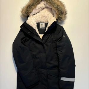 Svart vinterjacka med fuskpäls - Säljer en svart vinterjacka från McKinley i mycket bra skick. Jackan har en mysig fuskpäls runt huvan och är fodrad med fleece för extra värme. Den har långa ärmar och reflexdetaljer för ökad synlighet. Perfekt för kalla vinterdagar.