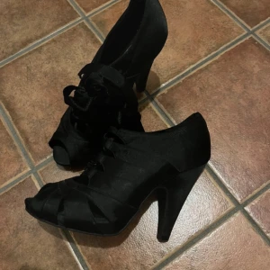 Svarta klackskor från Din Sko - Snygga svarta klackskor från Din Sko i bra skick. De har en elegant snörning och en öppen tå-design som ger en chic look. Perfekta för festliga tillfällen eller en utekväll. Klackarna ger en fin höjd och är bekväma att gå i.