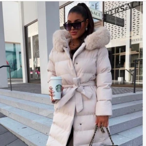 Beige dunjacka med pälskrage - Säljer en superfin beige dunjacka i storlek M. Den har en mysig pälskrage och ett bälte i midjan för en snygg passform. Perfekt för kalla vinterdagar! Jackan är i nytt skick och har både knappar och dragkedja framtill.