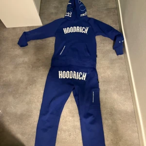 Blå hoodie från Hoodrich - Säljer en snygg blå hoodie från Hoodrich. Den har en stor logga på bröstet och coola detaljer på ärmarna. Perfekt för en avslappnad stil eller streetwear-look. Passar bra till både vardag och chill.