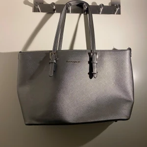 Grå axelväska från Michael Kors - Snygg och stilren grå axelväska från Michael Kors i syntetmaterial. Väskan har en praktisk dragkedja på baksidan och justerbara remmar. Perfekt för både vardag och fest!