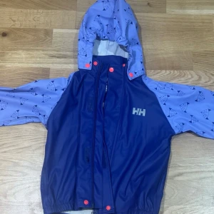 Blå regnjacka från Helly Hansen - Säljer en blå regnjacka från Helly Hansen i mycket bra skick. Jackan har ett coolt mönster med drakar och är perfekt för regniga dagar. Den har en praktisk huva och stängs med dragkedja och knappar. Perfekt för höst och vår! Täckbyxa medföljes!