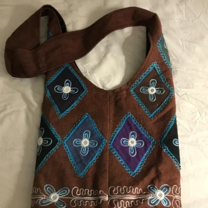 Brun axelväska med broderade mönster - Säljer en unik brun axelväska med färgglada broderade mönster i blått, lila och vitt. Säljs då den tyvärr inte kommit till användning 