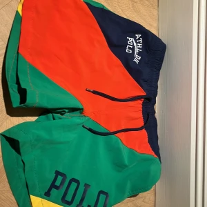Färgglada simshorts från Ralph Lauren - Säljer ett par färgglada simshorts från Ralph Lauren i storlek M barnstorlek. De har en cool mix av grönt, orange och blått med 'POLO' tryckt på sidan. Shortsen har en elastisk midja med snörning och en praktisk ficka med knapp. Perfekta för sommaren! 🌞ALDIRG ANVÄNTS!!