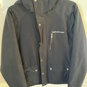 Svart jacka från Peak Performance - Säljer en svart jacka från Peak Performance i storlek L. Den har en dragkedja och knappar framtill, samt en praktisk bröstficka med dragkedja. 