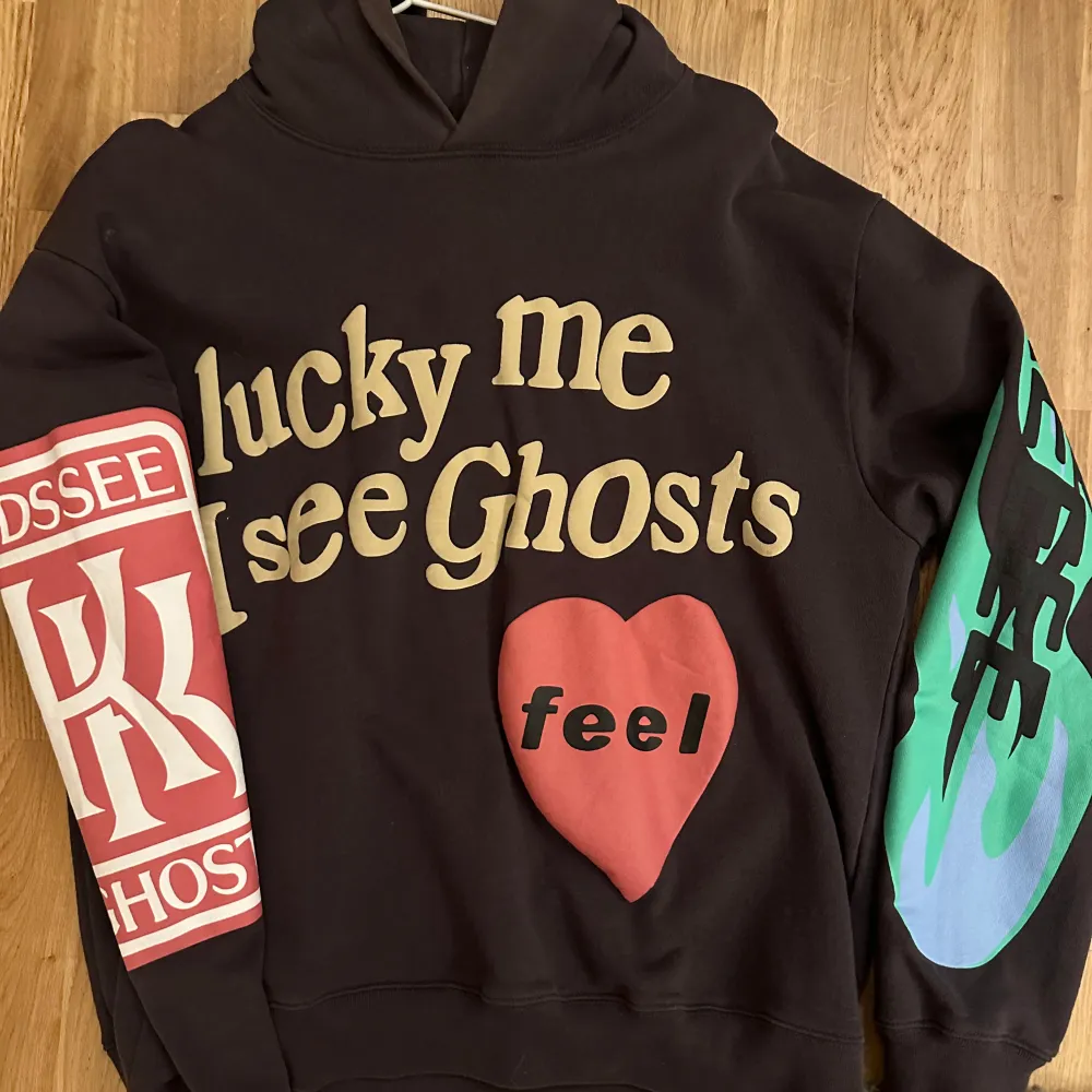 Säljer en hoodie med coolt tryck 'lucky me I see Ghosts' och ett rött hjärta med texten 'feel'. Ena ärmen har ett rött tryck med bokstäver och den andra har ett grönt och blått mönster. Perfekt för höst och vinter!. Hoodies.