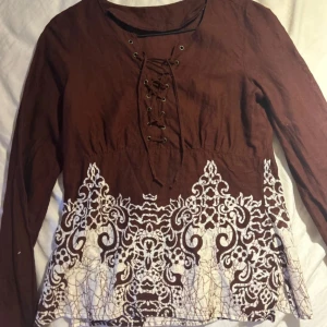 Brun långärmad topp med spetsdetaljer - Säljer en superfin brun långärmad topp med snygga vita spetsdetaljer längst ner. Den har en cool snörning framtill som ger en unik touch. 