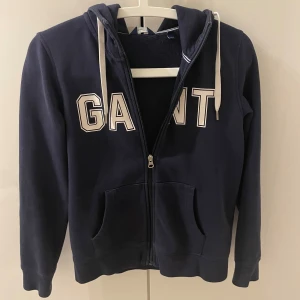 Mörkblå hoodie från GANT - Säljer en snygg mörkblå hoodie från GANT i storlek XS. Den har en dragkedja framtill och stora bokstäver på bröstet. Perfekt för höst och vår, med en skön passform och praktiska fickor. Huvan har vita snören för justering. Passar både till vardags och chillkvällar. Säljer pågrund av att den inte kommer till användning. Köpt från dam men passar killar också.