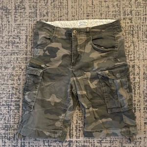 Camouflage shorts från Jack & Jones - Säljer ett par snygga camouflage shorts från Jack & Jones i bra skick. De har flera praktiska fickor med dragkedja och knappar, perfekt för sommarens äventyr. Materialet är slitstarkt och bekvämt, idealiskt för både vardag och utomhusaktiviteter.