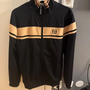 Svart träningsset från Sergio Tacchini - Säljer ett snyggt svart träningsset från Sergio Tacchini. Jackan har en dragkedja och beige ränder över bröstet och ärmarna. Byxorna har en bekväm passform med dragsko i midjan och matchande logga. Perfekt för både träning och chill. Skick 10/10 