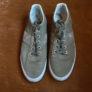 Gröna sneakers från Polo Ralph Lauren - Säljer ett par gröna sneakers från Polo Ralph Lauren i bra skick. Skorna har en klassisk design med snörning och broderad logga på sidan. De är tillverkade i mocka och syntetmaterial med en bekväm sula. Perfekta för vardagsbruk och ger en stilren look.