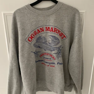 Grå sweatshirt med Ocean Market-tryck - Säljer en grå sweatshirt med ett coolt Ocean Market-tryck i rött och blått. Tröjan har en avslappnad passform och är perfekt för vardagsbruk. Den är i bra skick och superbekväm att ha på sig. Perfekt för höst och vår!