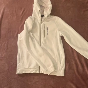 Vit hoodie från Sail Racing - Säljer en snygg vit hoodie från Sail Racing i storlek 176. Den har en dragkedja framtill och en praktisk bröstficka med dragkedja. Perfekt för vår och höst, med en stilren design och bekväm passform. Huvan ger extra skydd mot kyliga vindar. Passar både till vardags och lättare aktiviteter. Bra skick då den endast är använd ett par gånger, Nypris: 999 kr
