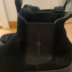 Svarta boots från Canada Snow - Snygga svarta boots i mocka från Canada Snow. De har en stilren design med elastiska paneler på sidorna för enkel på- och avtagning. Perfekta för höst och vinter med en bekväm passform och robust sula.