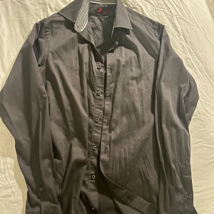 Grå slim fit skjorta - Snygg grå skjorta från Olymp i slim fit-modell. Den har långa ärmar och en stilren design med knappar framtill. Kragen har ett randigt mönster i svart och vitt som ger en extra touch. Perfekt för både vardag och fest!