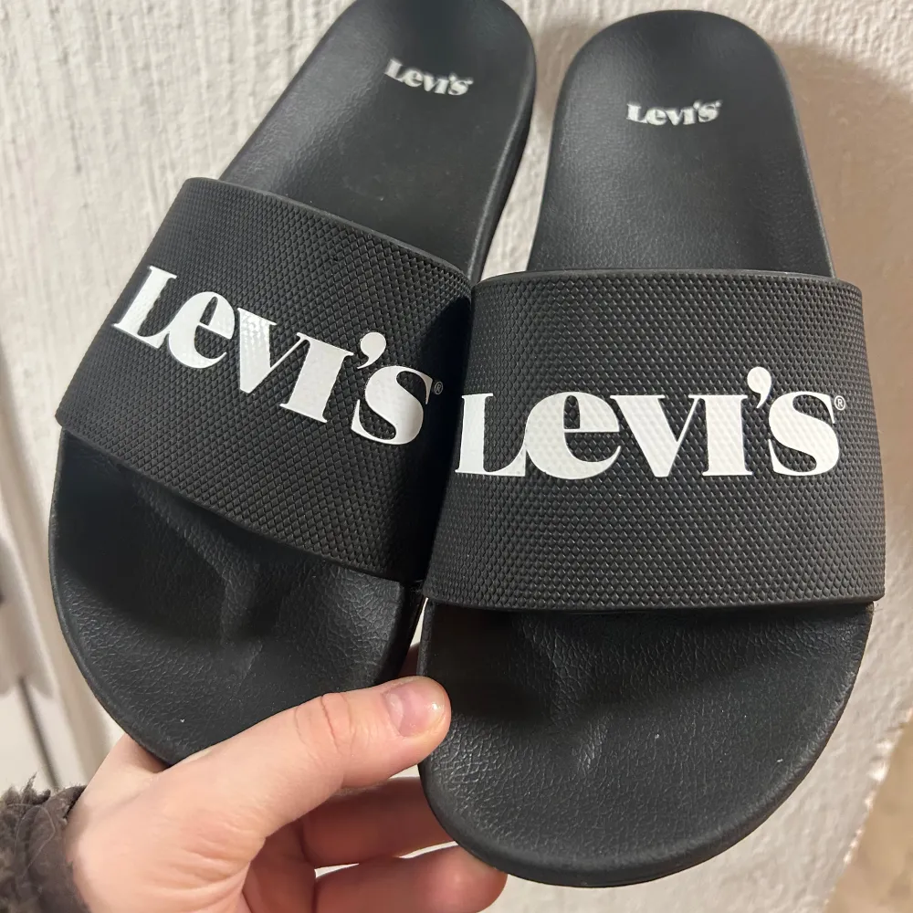 Säljer ett par svarta sandaler från Levi's i nytt skick. De har en bred rem med vit logotyp och är perfekta för sommaren. Superbekväma och stilrena för både stranden och stan!. Skor.