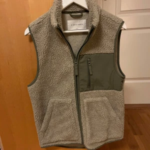 Grön/beige fleeceväst från A Day's March - Säljer en supermysig grå fleeceväst från A Day's March. Den har en dragkedja framtill och praktiska fickor. Perfekt för höst och vår när du vill hålla dig varm men ändå se stilren ut. Västen har en normal passform och är i mycket bra skick.