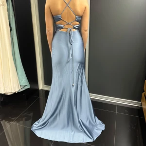 Ljusblå långklänning med öppen rygg - Elegant ljusblå långklänning i satin med en vacker öppen rygg och smala axelband som korsas. Klänningen har en smickrande omlott-design framtill och en slits för extra rörelsefrihet. Perfekt för festliga tillfällen!