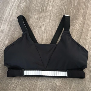 Svart sporttopp från Nike - Säljer en svart sporttopp från Nike i mycket bra skick. Toppen har justerbara axelband och en vit detalj på ryggen. Perfekt för träning och ger bra stöd. Den är tillverkad i ett syntetmaterial som andas.